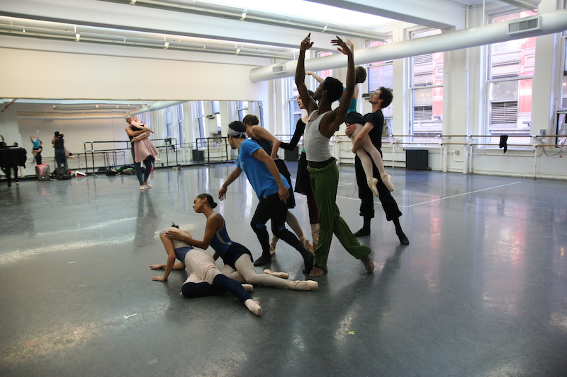 The cast of New Notes during a studio rehearsal. They pose in a tableau with some dancers sitting on the floor while others stand. A couple dancing is in the background. The dancers wear practice clothes.